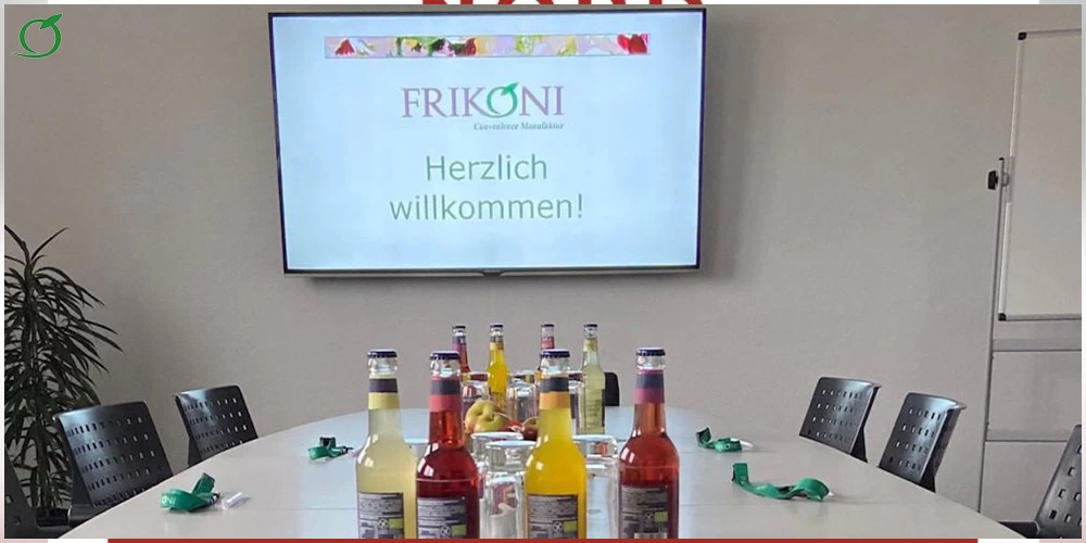 Bild zeigt einen Besprechungsraum mit einem Besprechungstisch mit sechs Stühlen im Vordergrund, auf dem sich verschiedene Getränkeflaschen befinden. Auf der Wand dahinter ein Monitor mit dem FRIKON-Logo und dem Text "Herzlich willkommen!"